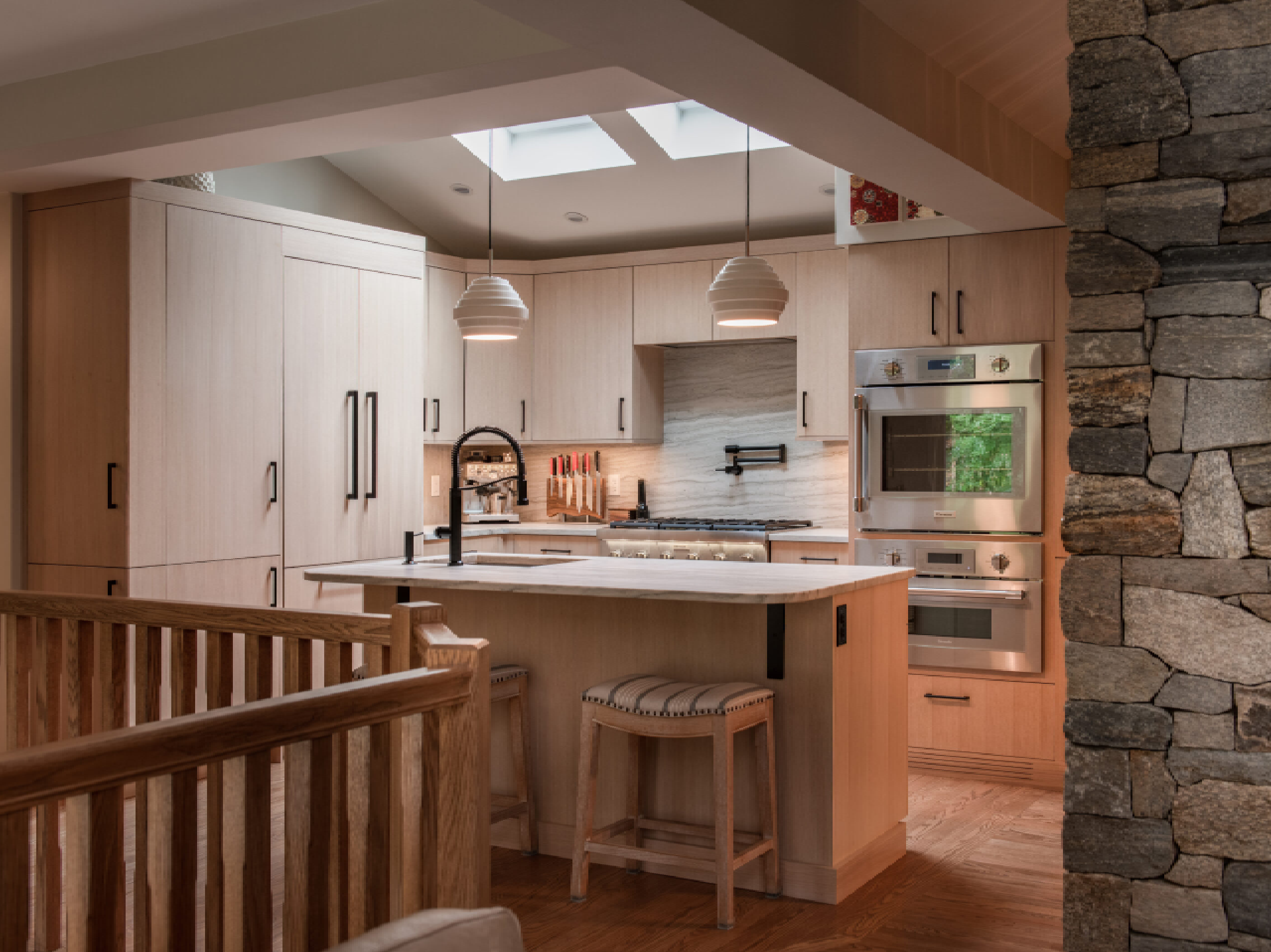 A cozy, modern kitchen with light wood cabinetry and stone accents. features include a central island, contemporary appliances, and pendant lighting, all within a warmly lit interior.